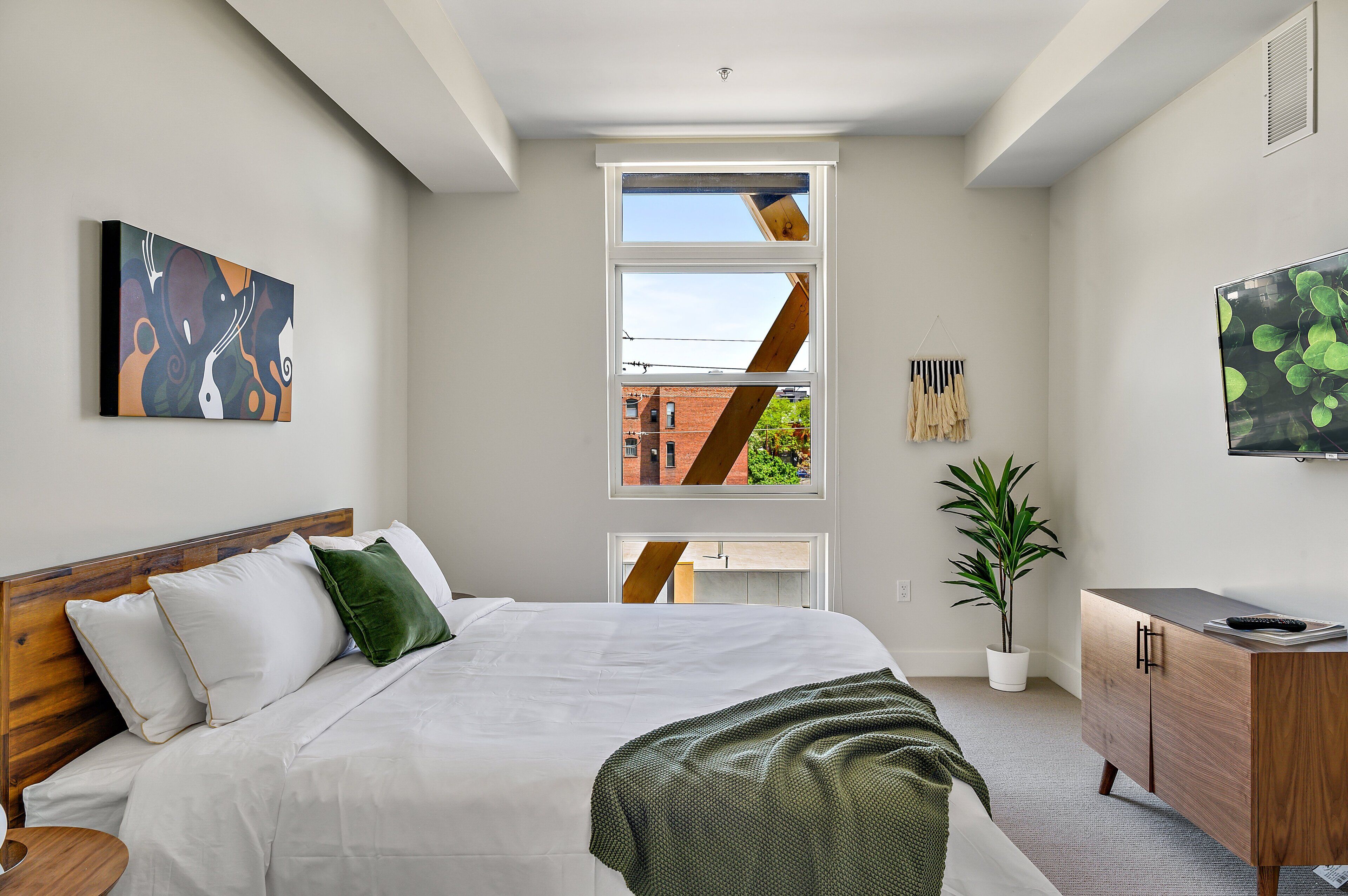 Een moderne slaapkamer met een groot bed, een televisie, een plant en een raam met uitzicht op een bakstenen gebouw.
