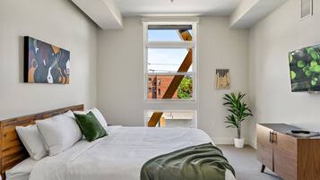 Een moderne slaapkamer met een groot bed, een televisie, een plant en een raam met uitzicht op een bakstenen gebouw.