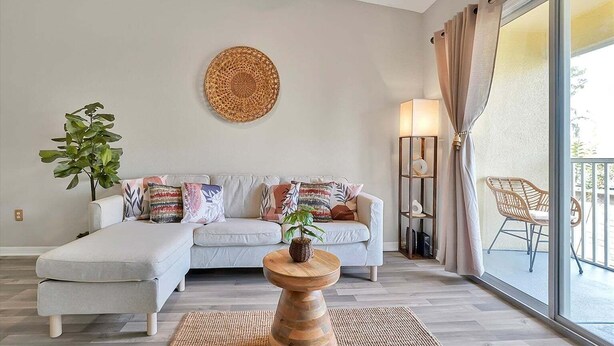 A living room with a sofa, cushions, a round wall decoration, a floor lamp, a side table with a plant, and a view of the outside through a sliding door. - 2722 Club Cortile Unit B 3bed (Kissimmee)