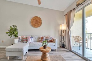 A living room with a sofa, cushions, a round wall decoration, a floor lamp, a side table with a plant, and a view of the outside through a sliding door. - 2722 Club Cortile Unit B 3bed (Kissimmee)