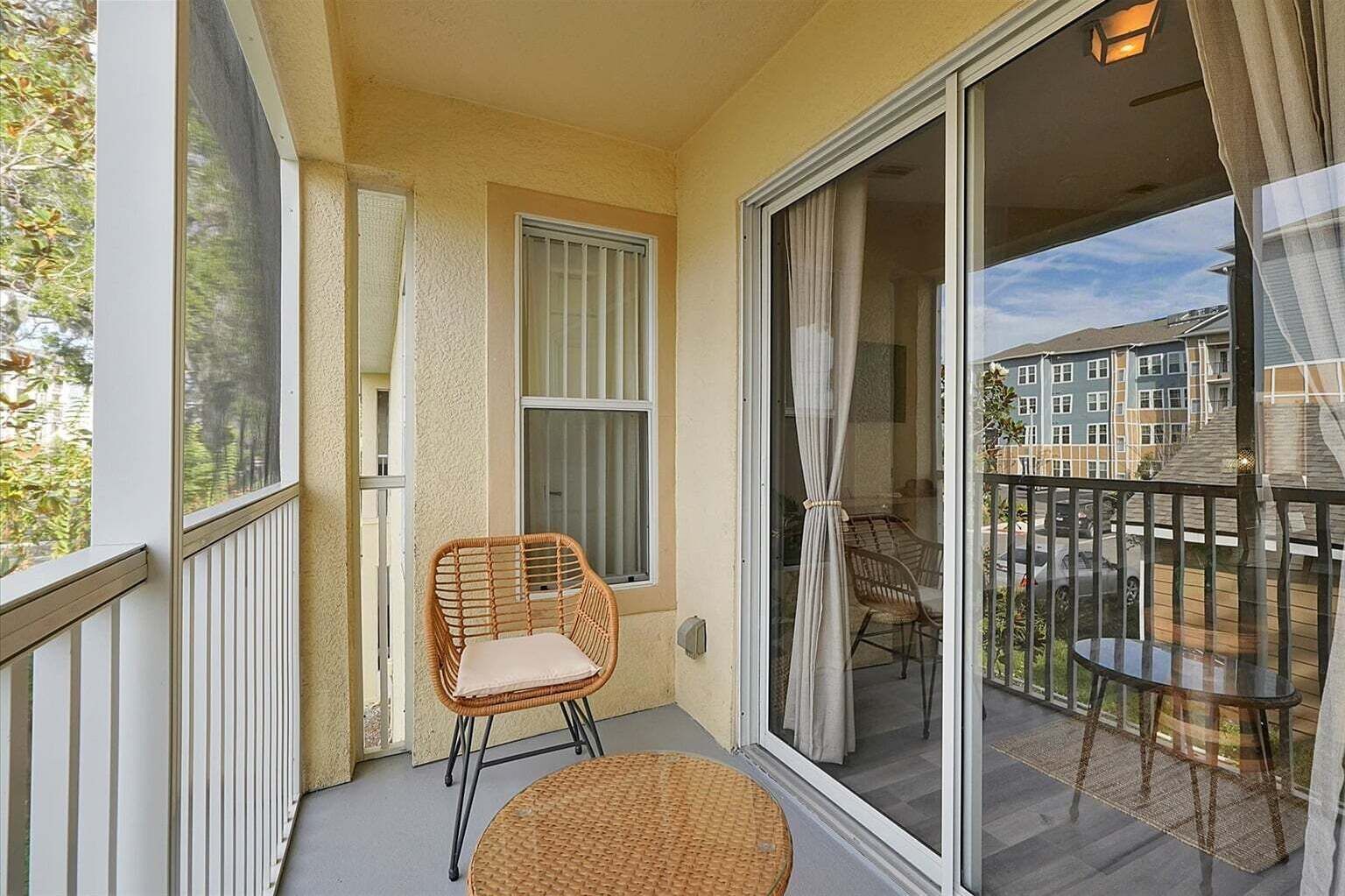 Un balcon avec un fauteuil en osier et une petite table, une porte-fenêtre coulissante et une vue sur un quartier résidentiel.