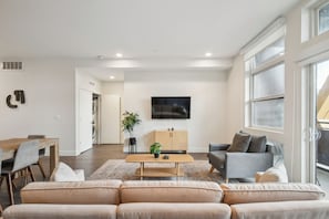 En moderne stue med sofa, sofabord og fjernsyn. Der er et spiseområde med bord og stole, og et køkken i baggrunden.
