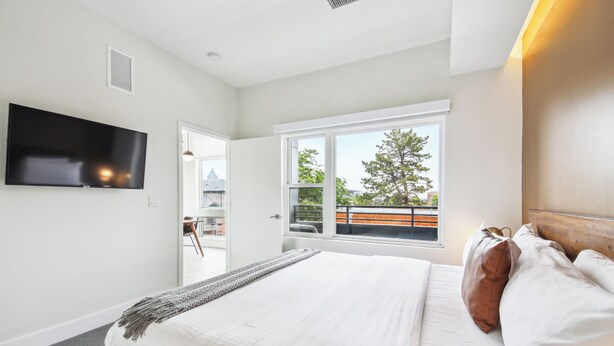 Un dormitorio con una cama grande, un televisor montado en la pared, una ventana con vistas al exterior y una puerta que da a otra habitación.
