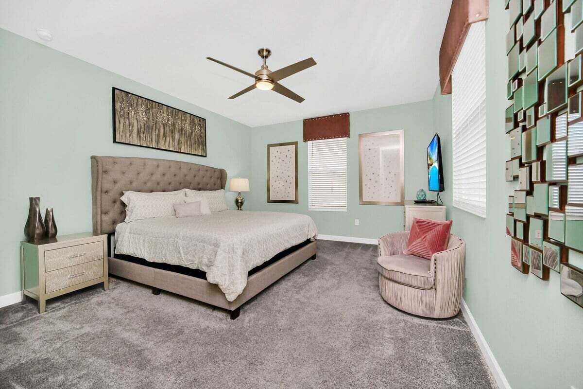 A bedroom with a tufted headboard bed, a bedside table, a chair, a ceiling fan, and a wall with framed pictures.