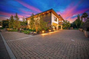 Une maison moderne à deux étages, avec une allée en briques, entourée d’une végétation luxuriante et de palmiers.