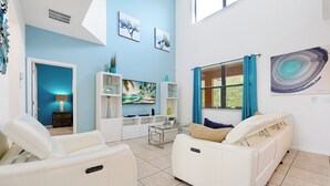 A modern living room with white leather sofas, a glass coffee table, and a flat-screen TV. There are shelves with decorative items and a painting on the wall. - 2616 Tranquility, Kissimmee, 6 BED (Kissimmee)