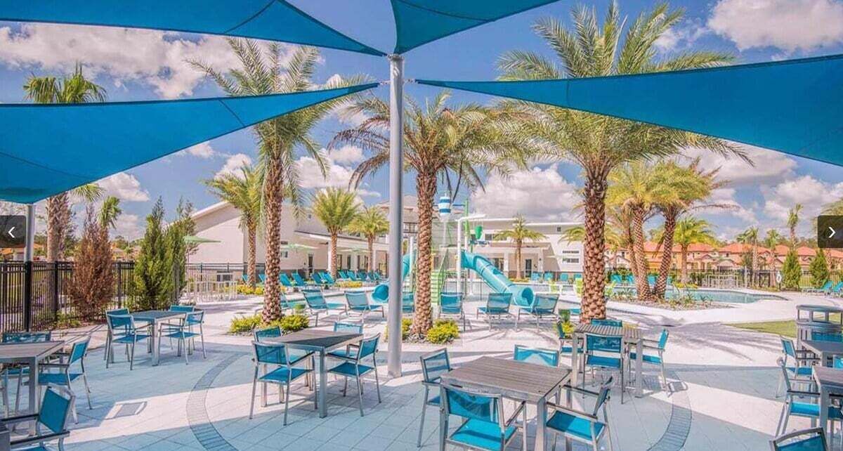 An outdoor seating area with tables and chairs, palm trees, and a pool in the background.