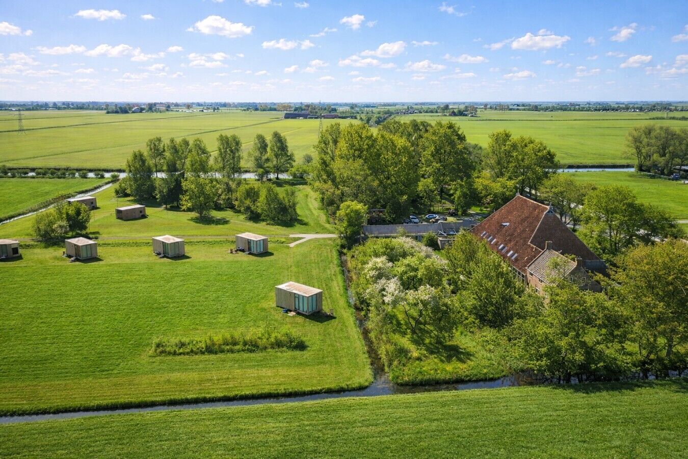 Un paysage rural avec une grande maison et plusieurs petits bâtiments, entourés de champs verdoyants et d’arbres.