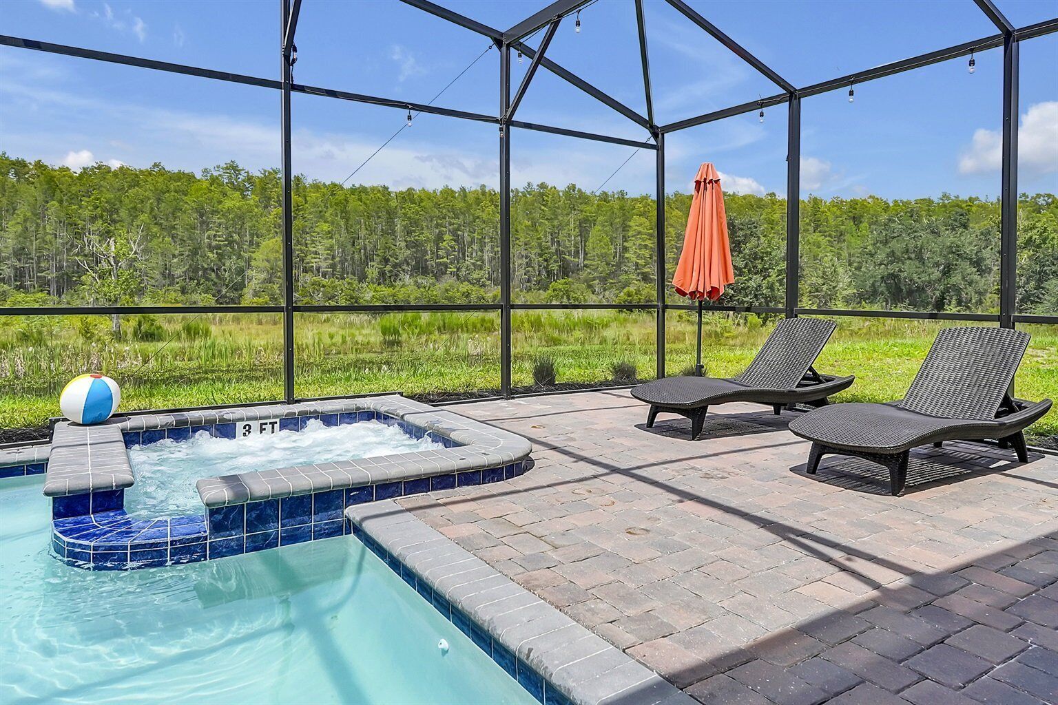 A hot tub with two lounge chairs and an umbrella, surrounded by a glass enclosure with a view of a forest.