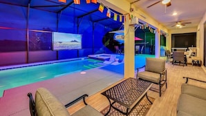 A pool area with a ceiling fan, seating, and a TV screen displaying a landscape. - 2473 Shanti · Deepsea World/15 Suites/sleep 30/oversize Pool&spa (Kissimmee)