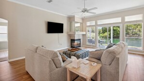A living room with a fireplace, a ceiling fan, and large windows. - 2888 Tidewater Drive (Fernandina Beach)