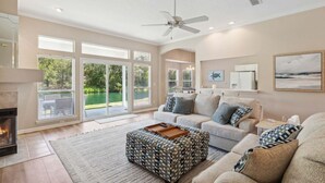 A spacious living room with a fireplace, large windows, a ceiling fan, and a patterned ottoman. - 2888 Tidewater Drive (Fernandina Beach)