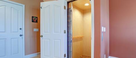 Un couloir avec deux portes blanches, dont l’une est ouverte et donne sur une salle de bain équipée d’une douche et d’un porte-serviettes.