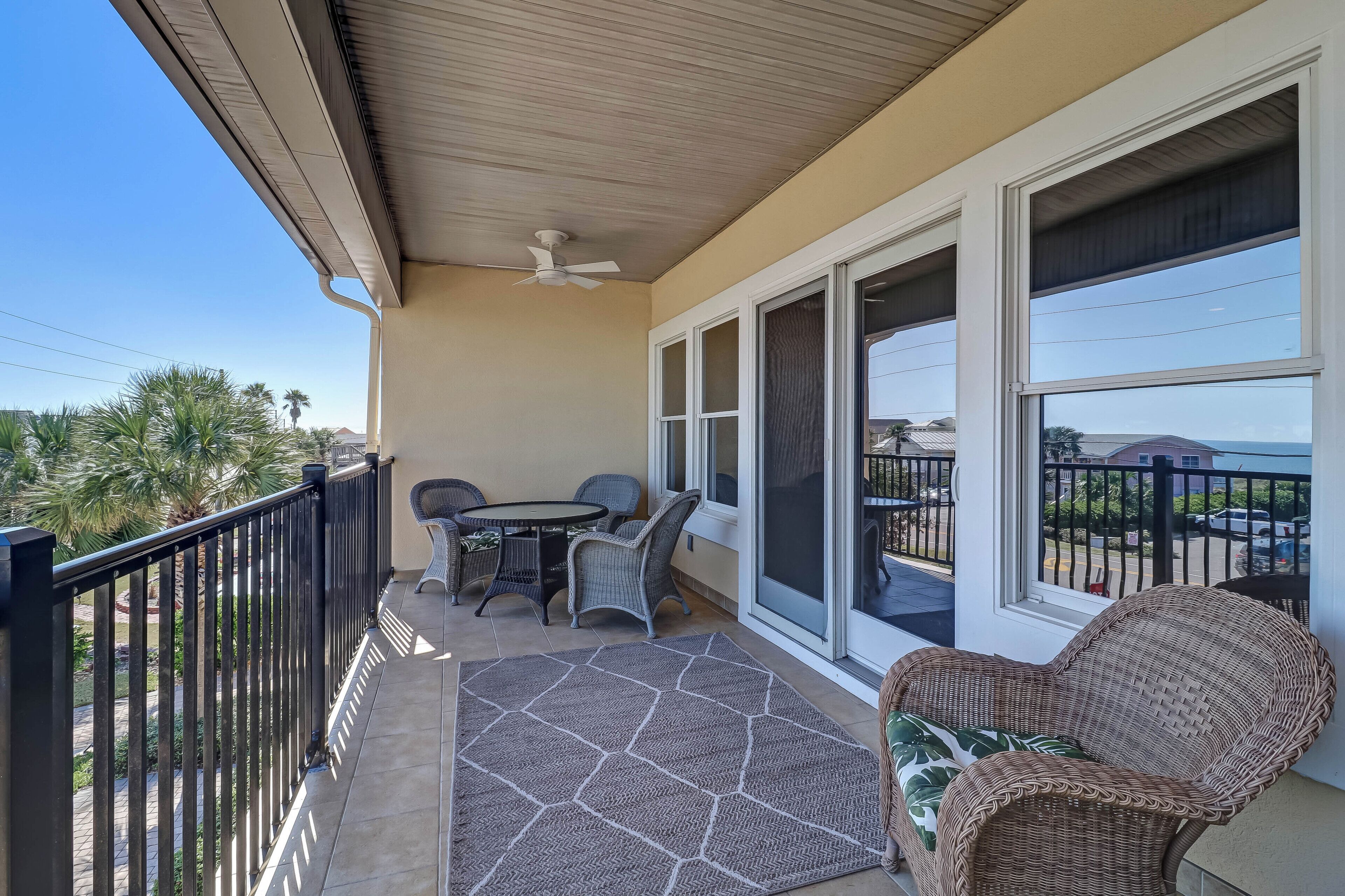 A balcony with a table and chairs, a wicker chair, and a view of the street and palm trees.