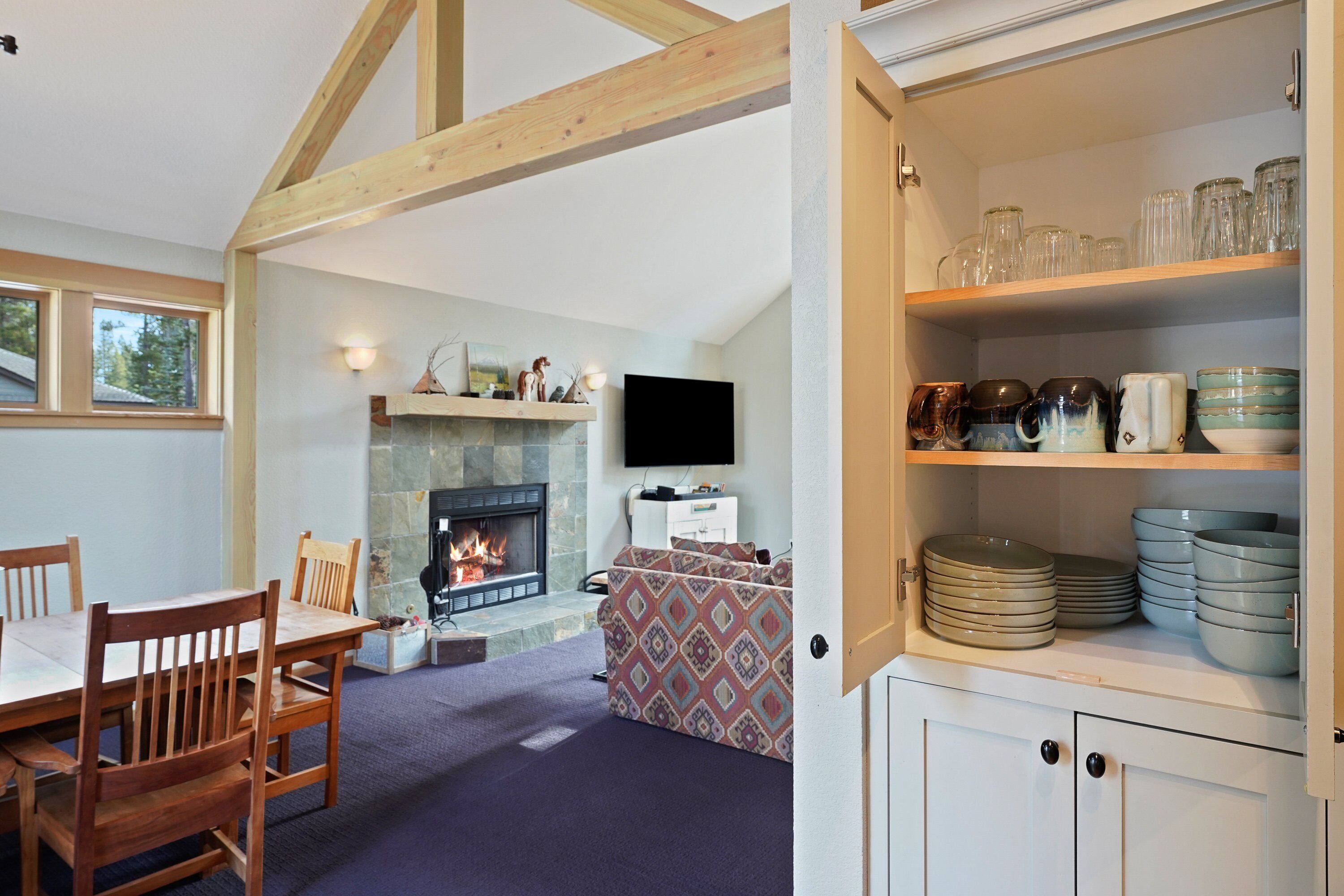 Ein gemütliches Wohnzimmer mit Kamin, einem Essbereich mit Holzsesseln und einem Regal mit Geschirr und Gläsern.