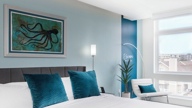 A modern hotel room with a large bed, teal pillows, a fluffy grey throw, a city view, and a wall art of an octopus. - 2880: Above the Cityline Elegance at Espadin (Denver)