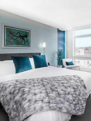 Una habitación de hotel moderna con una cama grande, almohadones color turquesa, un plaid gris suave y vistas a la ciudad, además de un cuadro de un pulpo.