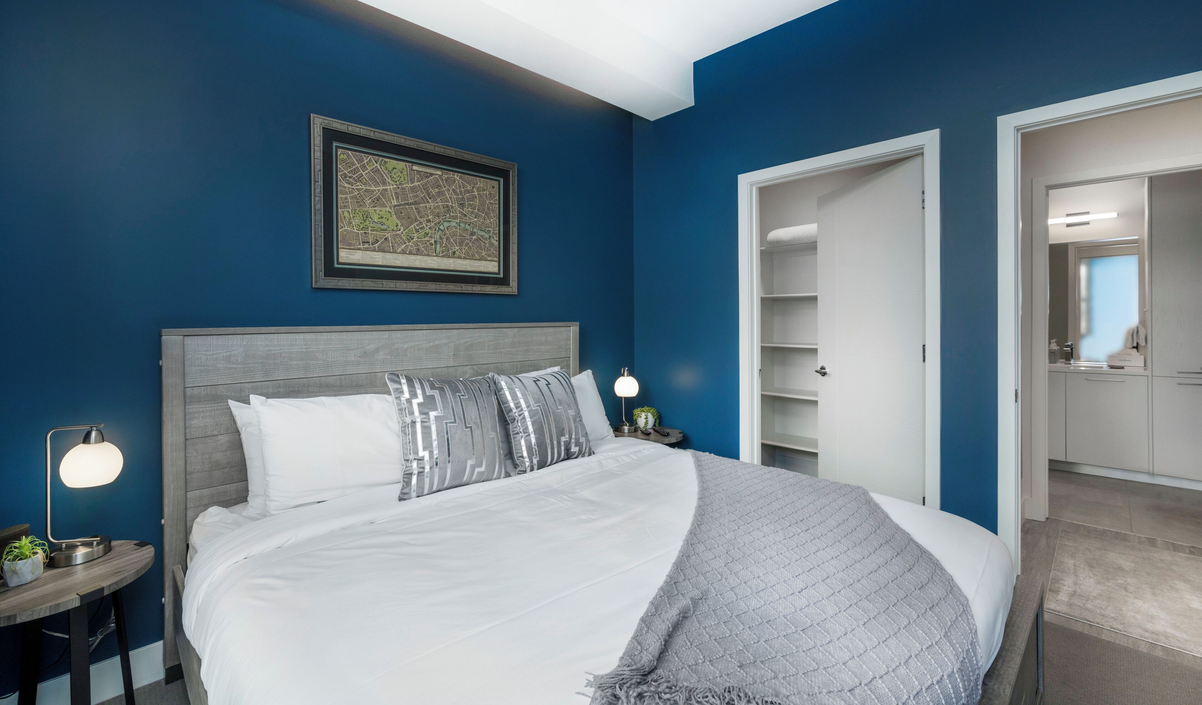 Een slaapkamer met een groot bed, een nachtkastje met een lamp, een inlijsting kaart aan de muur, en een kledingkast met planken.