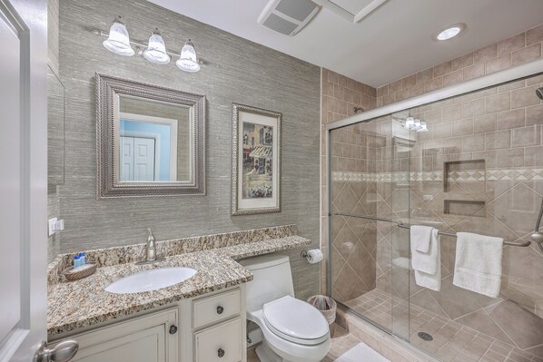A bathroom with a granite countertop sink, a toilet, a glass shower enclosure, and a mirror with three lights. - 2942 Atrium Villa (Seabrook Island)