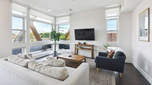 Ein geräumiges Wohnzimmer mit großem Fenster, einem Flachbildfernseher, einem Couchtisch aus Holz und einem dunkelblauen Sessel.