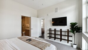 Ein modernes Schlafzimmer mit einem großen Bett, einem an der Wand montierten Fernseher, einer Topfpflanze und einem Holzregal.