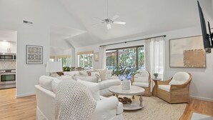 A spacious living room with a white sofa set, a round coffee table, and a wicker chair. There is a ceiling fan, a large window with curtains, and a kitchen area visible in the background. - 2908 Captain Sams (Seabrook Island)