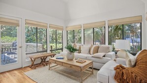 A bright, sunlit living room with a white sofa, a wooden coffee table, and a bench. Large windows offer views of greenery outside. - 2908 Captain Sams (Seabrook Island)