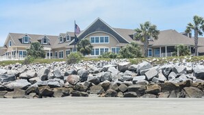 A large, multi-story house with a prominent American flag, surrounded by a rocky shoreline and palm trees. - 2908 Captain Sams (Seabrook Island)
