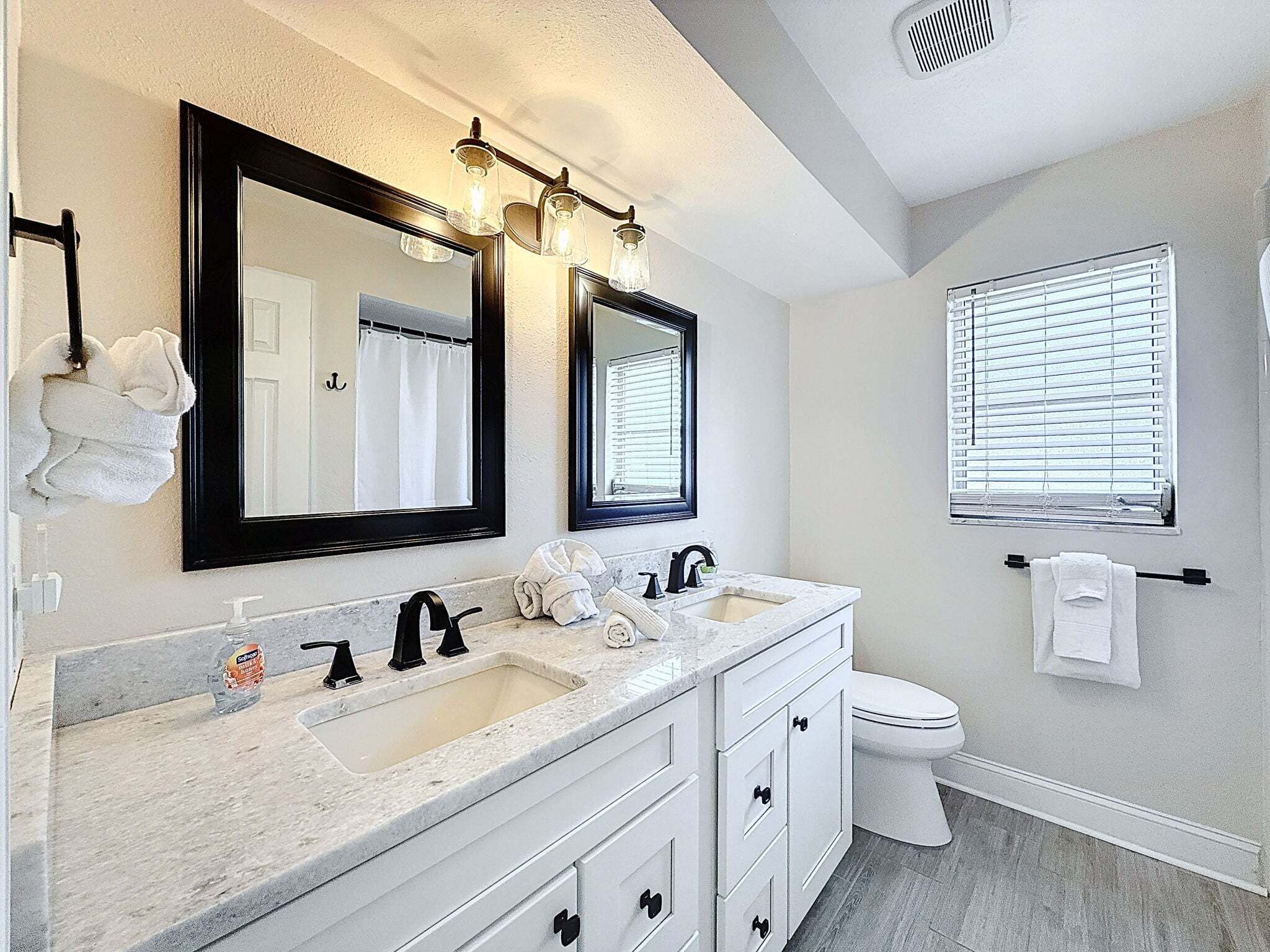 Une salle de bain avec deux lavabos, des toilettes et une fenêtre avec des stores.