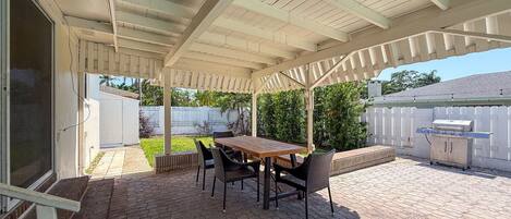 Un patio techado con suelo de ladrillo, una mesa de madera y sillas negras.