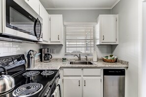 Una cocina con armarios blancos, estufa de acero inoxidable, microondas, lavavajillas y una ventana con persianas.
