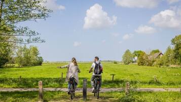 Twee personen op fietsen op een landweg, met groene velden en bomen, en een huis op de achtergrond.