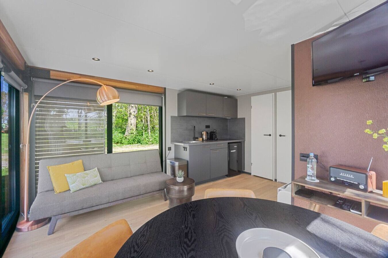 A modern living room with a sofa, a coffee table, and a dining area. There is a kitchen with grey cabinets and a dining table.