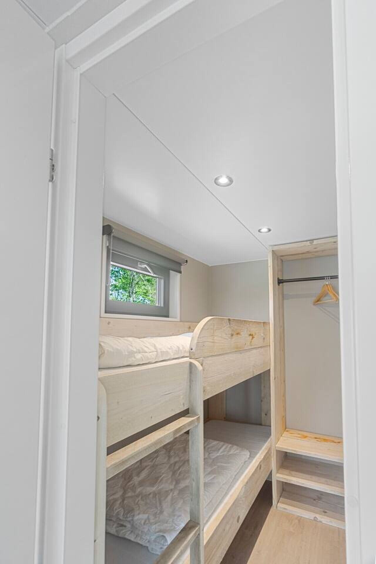 Une chambre avec un lit superposé, une fenêtre, une armoire en bois et un luminaire au plafond.