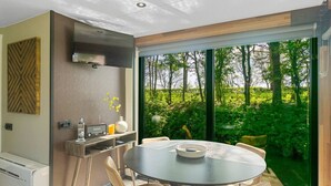 A modern kitchen with a built-in oven, a microwave, and a coffee machine. There is a dining table with chairs and a view of a lush garden through large windows.