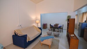 A living room with a blue sofa, a small ottoman, a wooden coffee table, a dining area with a table and chairs, and a kitchen area with a refrigerator and cabinets. - Rün Lirak, Lovely Home: Tremendous 2-bedroom house with AC in Guwahati (Guwahati)