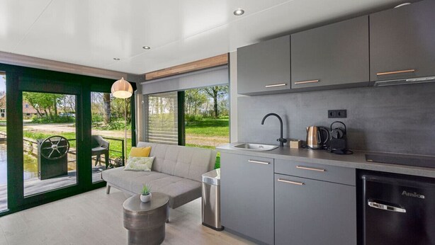 A modern kitchen with grey cabinets, a sink, and a coffee maker. There is a sofa and a small table with a plant. - Houseboat Doeke Sleat bij Leeuwarden (Weidum)