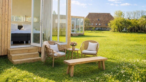 Een houten hutje met een glazen deur naar een slaapkamer, een tafeltje met bloemen, twee rieten stoelen en een bank.