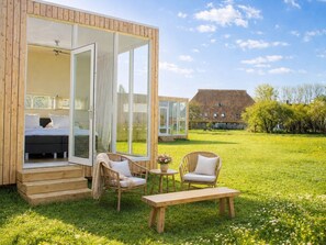 Eine Holzhütte mit Glastür zu einem Schlafzimmer, einem kleinen Tisch mit Blumen, zwei Korbstühlen und einer Bank.
