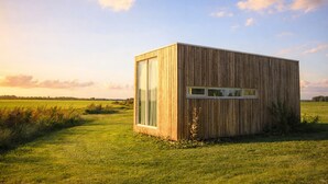 Een moderne houten blokhut met een groot raam, gelegen in een grasveld onder een heldere lucht.
