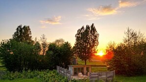 Een houten chalet met een terras en een enkele lounge fauteuil, omringd door een houten hek en bomen, met een zonsondergang op de achtergrond.
