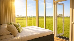 Een slaapkamer met een groot bed, uitzicht op een groen veld door een schuifdeur en een raam met gordijnen.