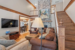 A spacious living room with a stone fireplace, a wooden staircase, and a television mounted on the wall.