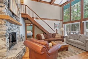 A spacious living room with a stone fireplace, leather sofas, a wooden staircase, and large windows offering a view of the outdoors.