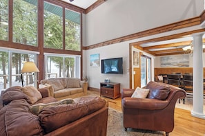 A spacious living room with large windows, a flat-screen TV, leather sofas, and a dining area with wooden furniture.