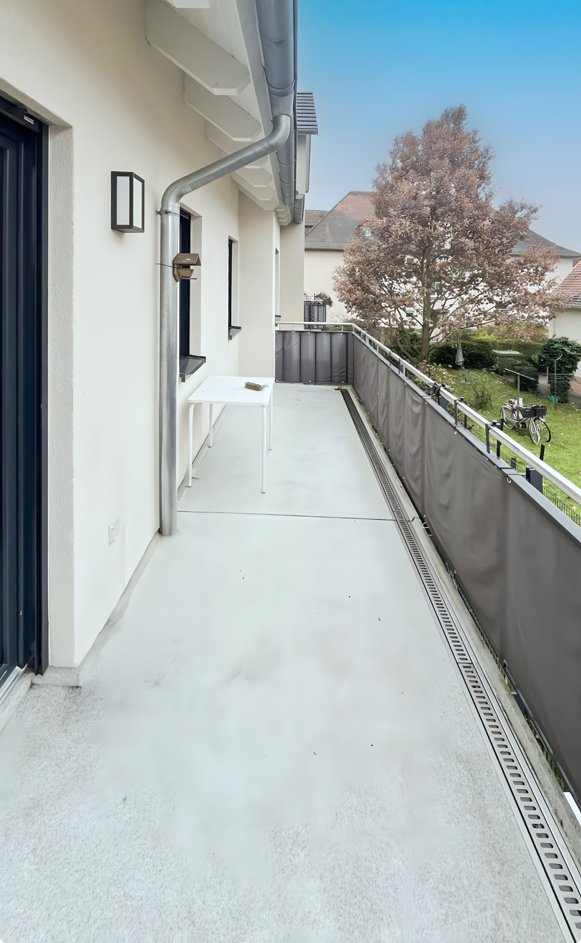 A balcony with a white table and chairs, a metal railing, and a view of a tree and a building in the background.