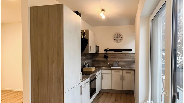 A modern kitchen with wooden flooring, white cabinetry, a built-in oven, and a large window. - Holiday Home 'Fortuna Suite' with Balcony and Wi-Fi (Wiesbaden)