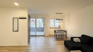 A modern living room with a black sofa, a dining area with a table and chairs, and a kitchen visible through an open door. - Holiday Home 'Fortuna Suite' with Balcony and Wi-Fi (Wiesbaden)