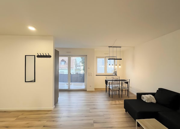 A modern living room with a black sofa, a dining area with a table and chairs, and a kitchen visible through an open door. - Holiday Home 'Fortuna Suite' with Balcony and Wi-Fi (Wiesbaden)