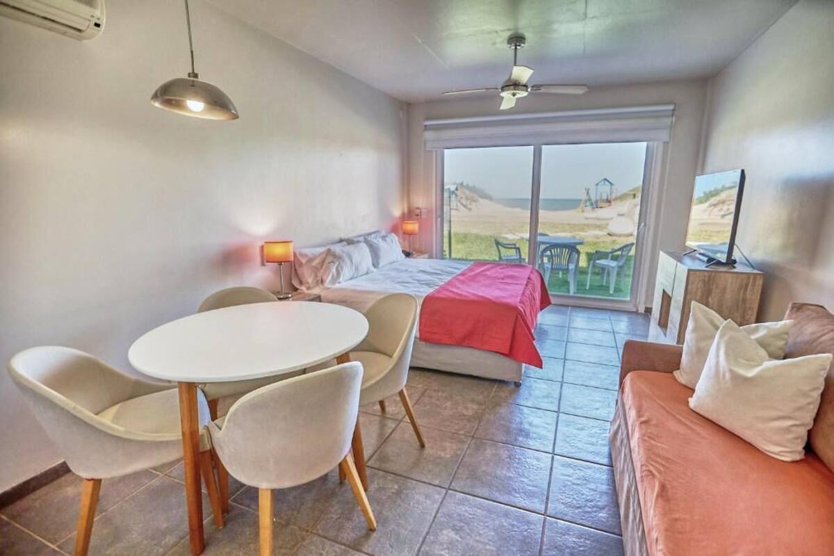 A room with a bed, a dining table with chairs, a sofa, and a TV. There is a large window with a view of the beach.